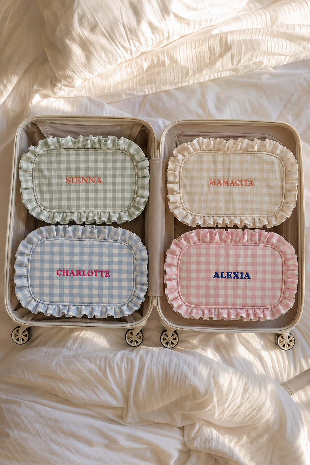 Four personalized luggage tags with ruffled edges are arranged inside an open beige suitcase on rumpled white bedding. The tags feature four different gingham patterns: sage green and white (labeled SURNA), cream and white (labeled MAMACITA), light blue and white (labeled CHARLOTTE), and pink and white (labeled ALEXIA).