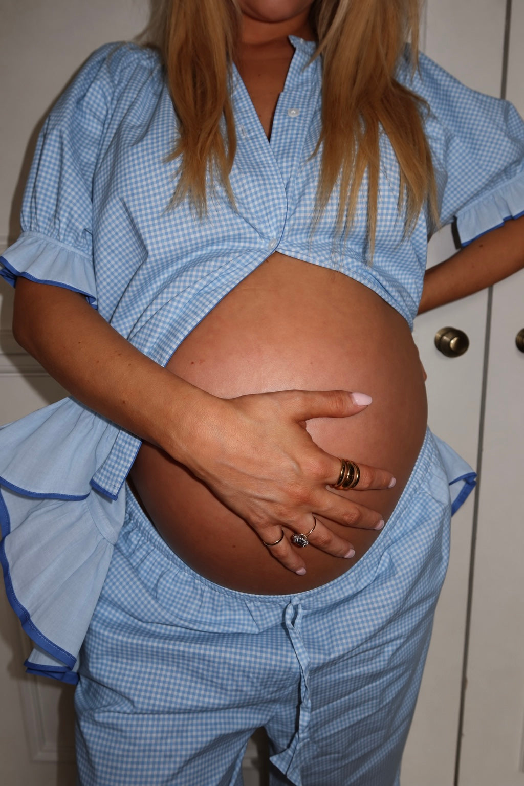A pregnant woman wearing a light blue and white gingham matching sleepwear set, with her top lifted to expose her belly, standing indoors.