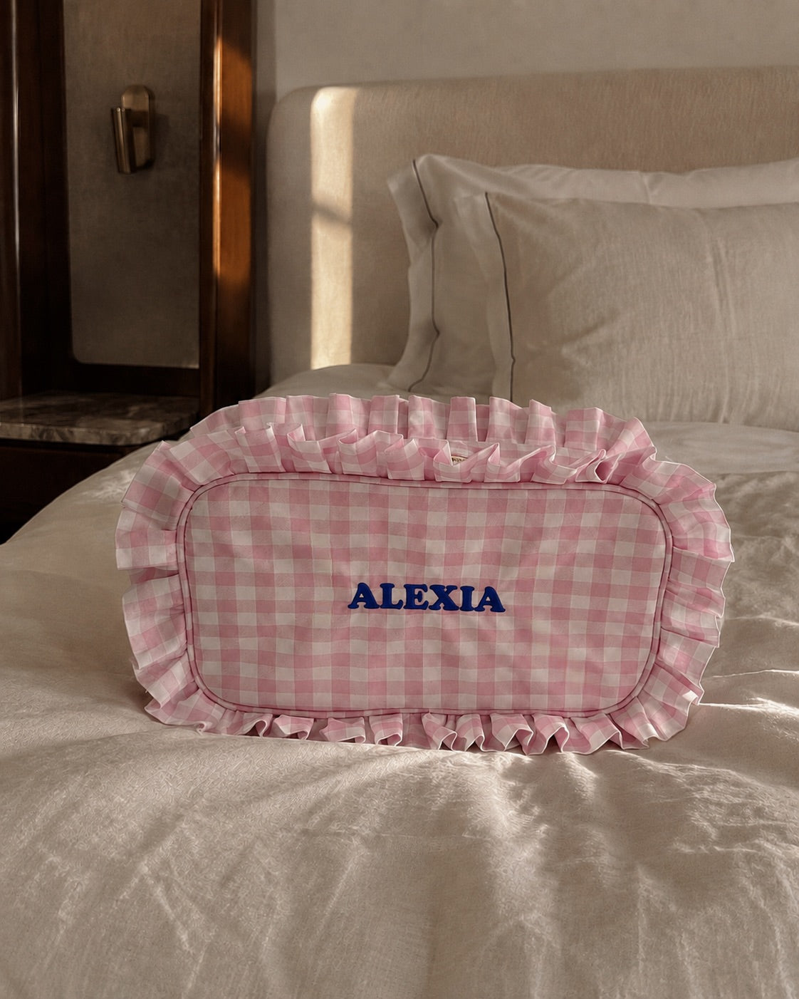 A pink and white gingham check ruffled pillow with the name 'ALEXIA' embroidered in blue on the front, resting on a beige linen bed in a softly lit bedroom.