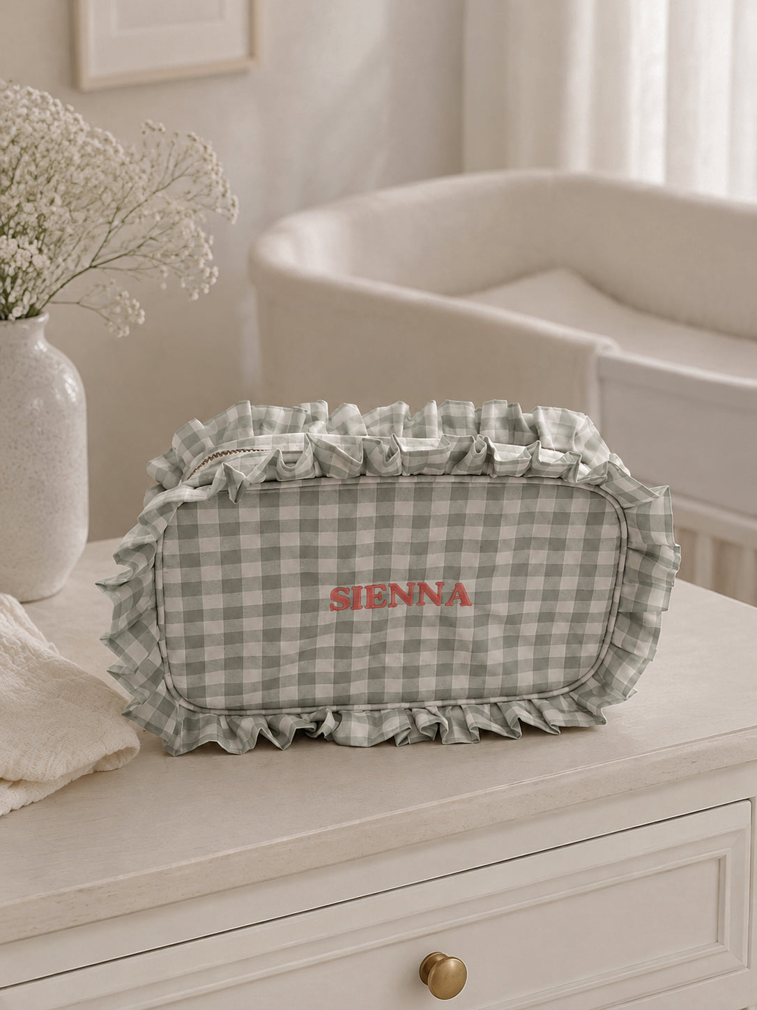A ruffled fabric cosmetic bag with a green and white gingham pattern and the brand name 'SIENNA' embroidered in red, displayed on a white dresser in a softly lit nursery setting.